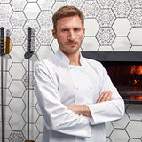 Longsleeve Chef Jacket in white worn by chef in kitchen setting with tiled wall and wood-fired oven, ideal for professional kitchenwear.